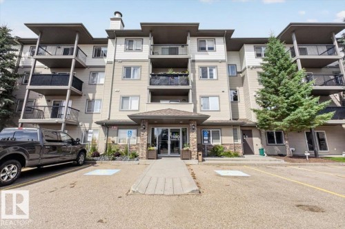 Multi-story building featuring balconies with black railings, a stone facade entrance, and mature evergreen landscaping - #421 1180 Hyndman Rd Nw, Edmonton, AB - Outdoor With Facade