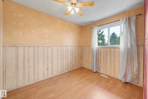 Spacious bedroom featuring warm golden-toned walls, beadboard wainscoting throughout, wood-finish flooring, a ceiling fan with integrated lighting, and large windows with natural light and garden views - 10032 173 Avenue Nw, Edmonton, AB - Indoor Photo Showing Other Room