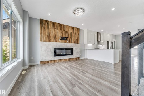 Open-concept living area featuring light wood-finish flooring, a modern electric fireplace with a patterned wood accent wall and light-toned tile surround, and recessed lighting - 6075 180 Avenue Nw, Edmonton, AB - Indoor Photo Showing Living Room With Fireplace