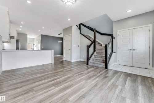 Open-concept living space with wood-finish flooring, a contemporary staircase featuring dark wood newel posts and glass panels, a kitchen with white cabinetry and a breakfast bar, recessed lighting, and a white double door entry - 6075 180 Avenue Nw, Edmonton, AB - Indoor