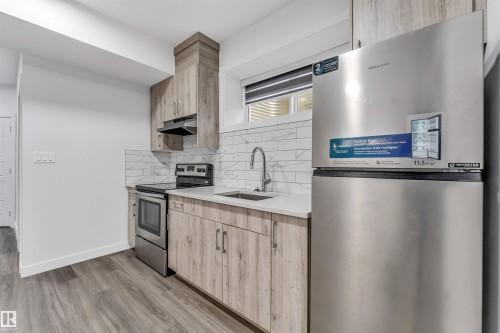 Compact kitchen featuring wood-finish cabinetry, stainless steel appliances, and a marble-look subway tile backsplash - 6075 180 Avenue Nw, Edmonton, AB - Indoor Photo Showing Kitchen With Stainless Steel Kitchen With Upgraded Kitchen