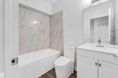 Bathroom featuring a white bathtub with marble-look tile surround, white vanity with integrated sink, silver-finish faucet, and wood-finish flooring - 6075 180 Avenue Nw, Edmonton, AB - Indoor Photo Showing Bathroom