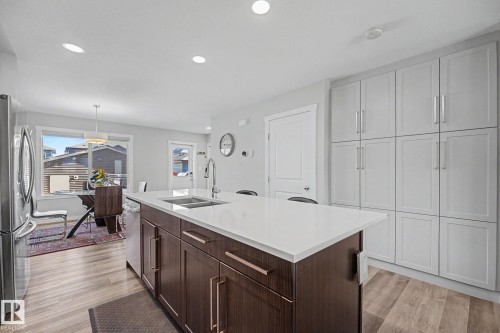 Kitchen featuring a large central island with a white countertop and dark wood-finish cabinetry, stainless steel sink with a gooseneck faucet, light wood-finish flooring, and extensive light gray built-in cabinetry with silver hardware - 2314 Wonnacott Crescent Sw, Edmonton, AB - Indoor Photo Showing Kitchen With Double Sink