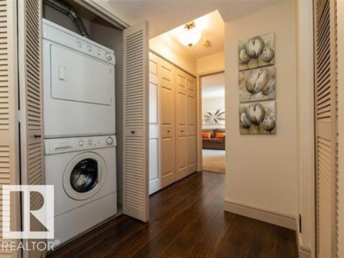 Dedicated laundry closet featuring a stacked white washer and dryer, louvered bi-fold doors, dark wood-finish flooring, and white baseboards - #1203 10025 113 St Nw, Edmonton, AB - Indoor Photo Showing Laundry Room