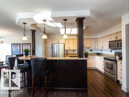 Kitchen featuring wood-finish flooring, light-toned cabinetry, stainless steel appliances, and a breakfast bar with pendant lighting - #1203 10025 113 St Nw, Edmonton, AB - Indoor Photo Showing Kitchen