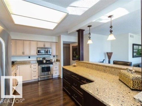 Kitchen featuring a large rectangular ceiling light panel, light-toned cabinetry, stainless steel appliances, speckled stone countertops, and wood-finish flooring - #1203 10025 113 St Nw, Edmonton, AB - Indoor Photo Showing Kitchen