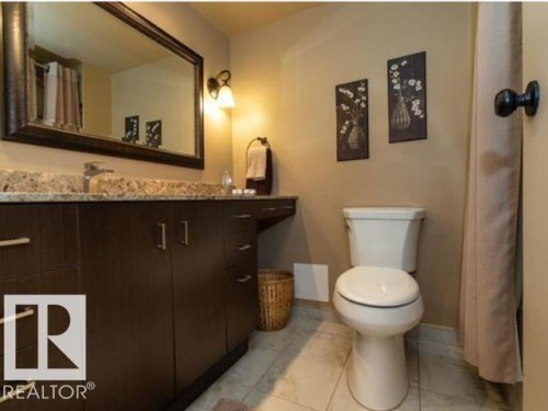 Bathroom featuring a dark wood-finish vanity with a stone-look countertop, a large framed mirror, a wall-mounted light fixture, and a white toilet - #1203 10025 113 St Nw, Edmonton, AB - Indoor Photo Showing Bathroom