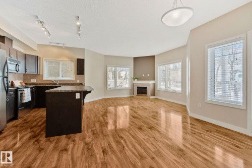 Open-concept living space featuring wood-finish flooring, a kitchen island with countertop seating, stainless steel appliances, and a corner fireplace with a mantel - #120 8931 156 St Nw, Edmonton, AB - Indoor Photo Showing Kitchen With Fireplace