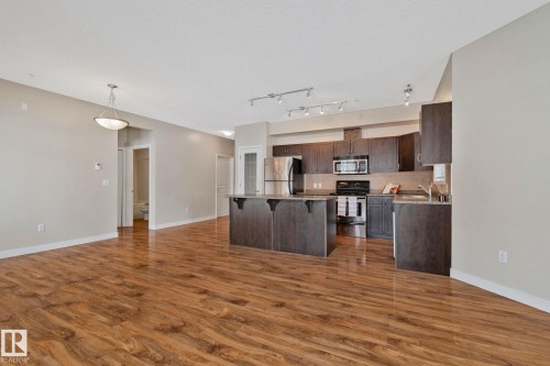 Open-concept living area featuring rich wood-finish flooring, a contemporary kitchen with dark wood cabinetry, stainless steel appliances, a kitchen island with seating, and track lighting - #120 8931 156 St Nw, Edmonton, AB - Indoor Photo Showing Kitchen