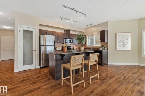 Open-concept kitchen featuring an island with seating, stainless steel appliances, dark wood cabinetry, and track lighting - #120 8931 156 St Nw, Edmonton, AB - Indoor Photo Showing Kitchen With Stainless Steel Kitchen With Upgraded Kitchen