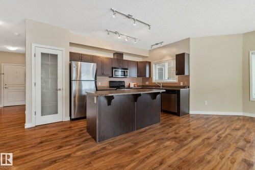 Open-concept kitchen and dining area featuring rich wood-finish flooring, dark cabinetry with stainless steel appliances, a substantial center island with seating for multiple guests, track lighting, and abundant counter and storage space - #120 8931 156 St Nw, Edmonton, AB - Indoor Photo Showing Kitchen With Stainless Steel Kitchen With Upgraded Kitchen