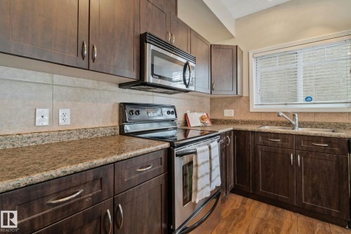 Elegant kitchen featuring rich wood-finish cabinetry with brushed metal hardware, granite countertops with decorative edge banding, and stainless steel appliances including a gas range and over-the-range microwave - #120 8931 156 St Nw, Edmonton, AB - Indoor Photo Showing Kitchen