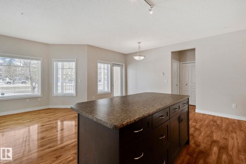 Spacious kitchen featuring a substantial dark-stained island with granite countertop and ample storage cabinetry, complemented by warm wood-finish flooring throughout - #120 8931 156 St Nw, Edmonton, AB - Indoor Photo Showing Other Room