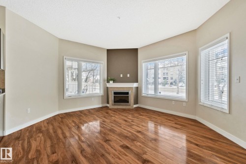 Open living area featuring wood-finish flooring, a corner fireplace with a mantel, and multiple large windows with blinds - #120 8931 156 St Nw, Edmonton, AB - Indoor Photo Showing Living Room With Fireplace
