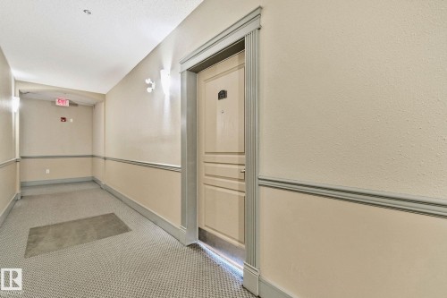 Hallway featuring textured walls, patterned carpeting, wall sconce lighting, and a detailed entry door with fluted trim - #120 8931 156 St Nw, Edmonton, AB - Indoor Photo Showing Other Room
