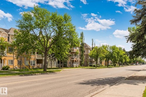 Street-level perspective showcasing apartment buildings with varied architectural styles, mature trees, and a clear sky with scattered clouds - #120 8931 156 St Nw, Edmonton, AB - Outdoor