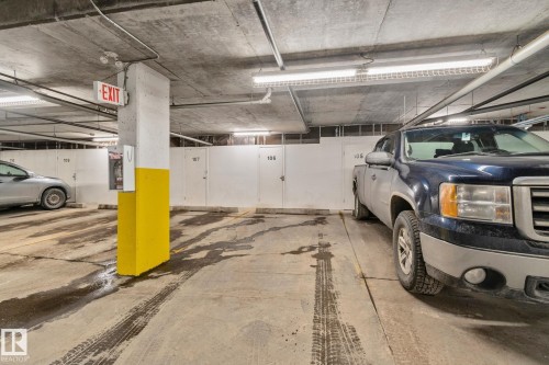 Underground parking space featuring a concrete floor and ceiling, fluorescent lighting, an exit sign, and individual storage units - #120 8931 156 St Nw, Edmonton, AB - Indoor Photo Showing Garage