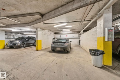 Secure, well-lit parking garage featuring concrete flooring, structural support columns with yellow safety striping, fluorescent ceiling lighting, and dedicated waste receptacle - #120 8931 156 St Nw, Edmonton, AB - Indoor Photo Showing Garage