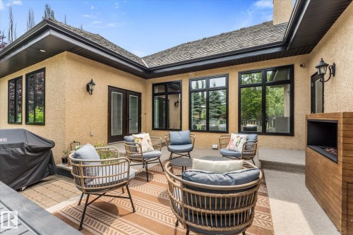 Spacious outdoor patio featuring textured stucco exterior, black-framed windows, and a built-in outdoor fireplace with wood-finish paneling - #52 Riverstone Close, Rural Sturgeon County, AB - Outdoor With Deck Patio Veranda With Exterior