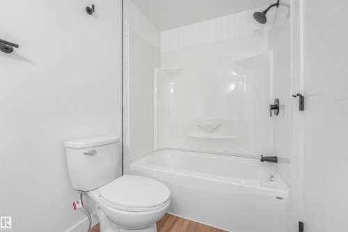Bathroom featuring a white bathtub and shower combination with dark-finished fixtures, a white toilet, and wood-finish flooring - #39 14511 52 Street Nw, Edmonton, AB - Indoor Photo Showing Bathroom