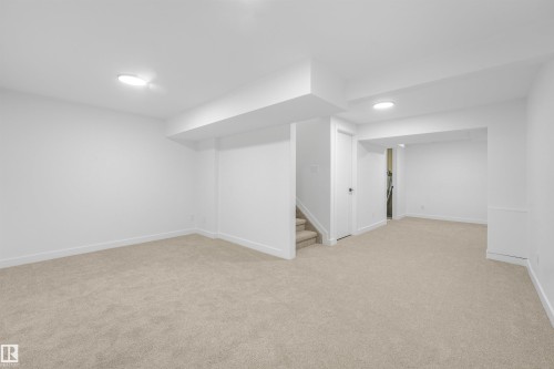 Spacious lower-level room featuring light-toned carpeting, crisp white walls, recessed ceiling lights, a white paneled door, and a carpeted staircase - #39 14511 52 Street Nw, Edmonton, AB - Indoor