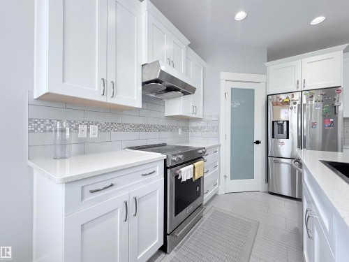 Kitchen featuring white shaker-style cabinetry, stainless steel appliances, quartz countertops, subway tile backsplash with mosaic accent, and a frosted glass pantry door - 8719 218 Street Nw, Edmonton, AB - Indoor Photo Showing Kitchen With Upgraded Kitchen