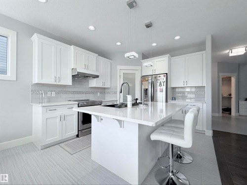 Modern kitchen featuring a large central island with seating, white shaker-style cabinetry, stainless steel appliances, recessed lighting, and tile flooring - 8719 218 Street Nw, Edmonton, AB - Indoor Photo Showing Kitchen With Upgraded Kitchen