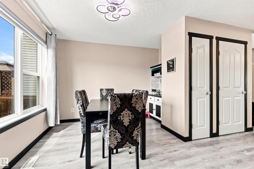 Modern kitchen and dining area featuring wood-finish flooring, a black upholstered dining chair with damask pattern, stainless steel appliances, white cabinetry, abundant natural light from large windows, and a contemporary ceiling fixture - 204 59 Street Sw, Edmonton, AB - Indoor Photo Showing Dining Room