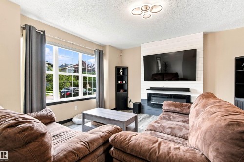 Spacious living room featuring multiple large windows with natural light, comfortable seating areas, wall-mounted flat-screen television above a fireplace with dark surround, built-in shelving, and hardwood-finish flooring throughout - 204 59 Street Sw, Edmonton, AB - Indoor Photo Showing Living Room With Fireplace