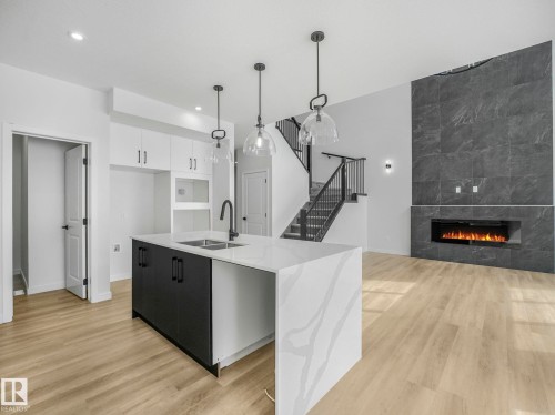Open-concept living area featuring a kitchen island with stone-finish countertop, wood-finish flooring, a contemporary fireplace, a stone-tiled accent wall, and a staircase with dark metal railings - 239 Kinglet Boulevard Nw, Edmonton, AB - Indoor Photo Showing Kitchen With Fireplace With Double Sink With Upgraded Kitchen