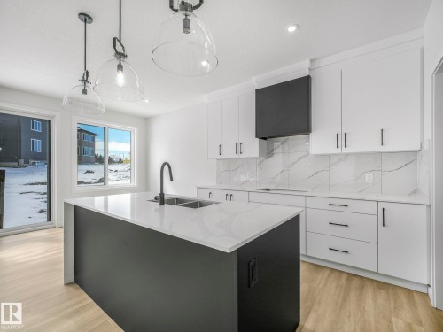 Modern kitchen featuring a dark gray island with a white countertop, light wood-finish flooring, white cabinetry with dark hardware, a patterned backsplash, and an integrated dark range hood - 239 Kinglet Boulevard Nw, Edmonton, AB - Indoor Photo Showing Kitchen With Double Sink With Upgraded Kitchen