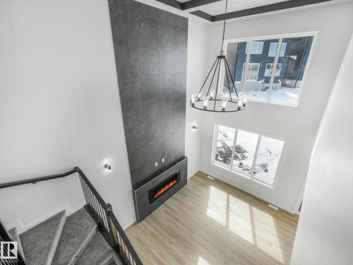 Two-story ceiling with a contemporary chandelier, floor-to-ceiling fireplace with an electric insert, large windows, wood-finish flooring, and a carpeted staircase with a metal railing - 239 Kinglet Boulevard Nw, Edmonton, AB - Indoor Photo Showing Other Room
