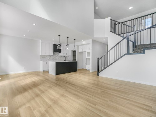 Open-concept living area featuring light wood-finish flooring, a modern kitchen island with an integrated sink, white cabinetry with dark hardware, a geometric backsplash, and a staircase with black metal railings - 239 Kinglet Boulevard Nw, Edmonton, AB - Indoor Photo Showing Kitchen