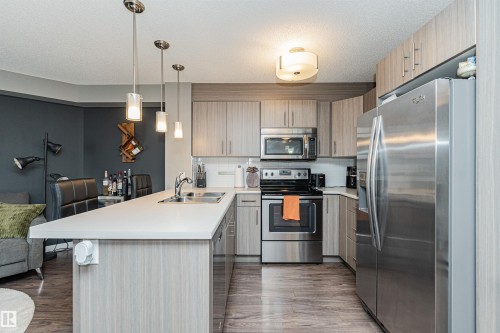 Modern kitchen featuring stainless steel appliances, white cabinetry with wood-finish accents, white quartz countertops, a large island with waterfall edge, pendant lighting, and wood-finish flooring throughout the open layout - #310 2229 44 Av Nw, Edmonton, AB - Indoor Photo Showing Kitchen With Double Sink With Upgraded Kitchen