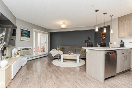 Open-concept living space with wood-finish flooring, kitchen island with stainless steel dishwasher, pendant lighting, and sliding glass door to outdoor area - #310 2229 44 Av Nw, Edmonton, AB - Indoor Photo Showing Kitchen
