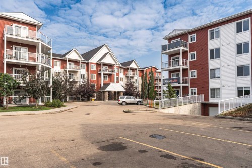 Multi-story building featuring red siding, white trim, multiple balconies with glass railings, and an entry canopy with stone accents - #310 2229 44 Av Nw, Edmonton, AB - Outdoor With Balcony With Facade