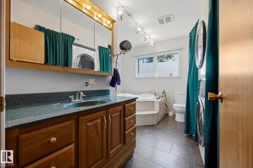 Spa-inspired primary bathroom featuring wood cabinetry with brushed brass hardware, dual vanity with dark stone countertop, recessed lighting, large framed mirror, and tile flooring - 9503 54 Street Nw, Edmonton, AB - Indoor Photo Showing Bathroom