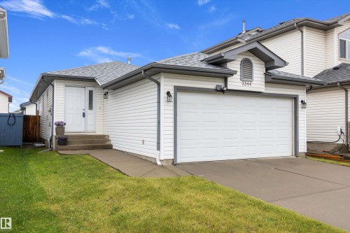 White siding exterior featuring an attached two-car garage, concrete driveway, front entry with a sidelight, and a peaked dormer accent - 3344 24 Avenue Nw, Edmonton, AB - Outdoor With Exterior