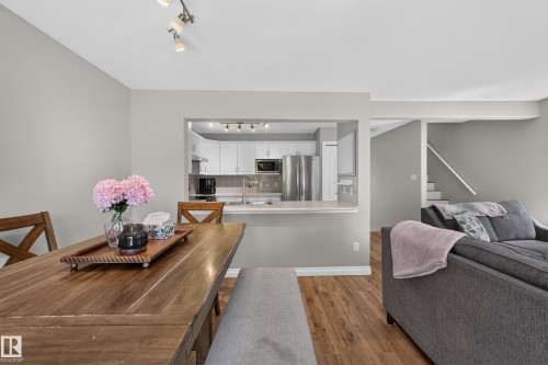 Open-concept living area showcasing warm wood-finish flooring throughout, a dining table with seating for multiple guests, and seamless sightlines to a modern kitchen featuring stainless steel appliances and white cabinetry - #25 200 Erin Ridge Dr, St. Albert, AB - Indoor Photo Showing Other Room