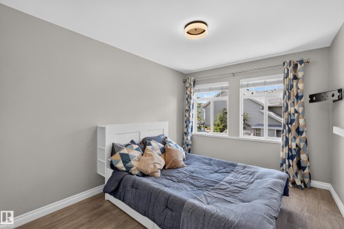 Bright secondary bedroom featuring a white bed frame, wood-finish flooring, large double-hung windows with patterned curtains allowing natural light, soft gray wall paint, and a recessed ceiling light fixture - #25 200 Erin Ridge Dr, St. Albert, AB - Indoor Photo Showing Bedroom