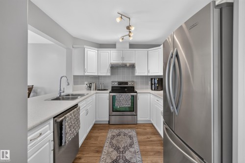 Kitchen featuring white cabinetry, stainless steel appliances, a double basin sink, light-toned countertops, and wood-finish flooring - #25 200 Erin Ridge Dr, St. Albert, AB - Indoor Photo Showing Kitchen With Double Sink With Upgraded Kitchen