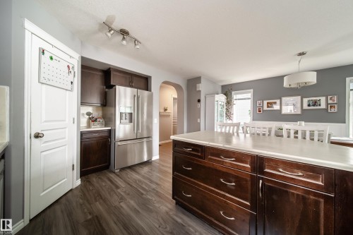 Wood-finish flooring throughout, dark wood-finish cabinetry, stainless steel refrigerator, kitchen island with a light-toned countertop, and recessed lighting - 7239 21 Avenue Sw, Edmonton, AB - Indoor Photo Showing Kitchen