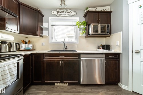 Thoughtfully appointed kitchen featuring dark wood cabinetry, stainless steel appliances including a dishwasher, undermount sink with chrome faucet, and neutral tile backsplash - 7239 21 Avenue Sw, Edmonton, AB - Indoor Photo Showing Kitchen