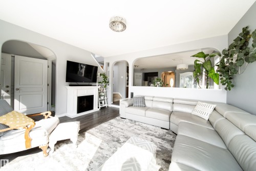 Bright open-concept living area featuring a curved sectional sofa, gas fireplace with white surround and mantel, wall-mounted flat-screen television, recessed ceiling lighting, and abundant natural light from multiple windows - 7239 21 Avenue Sw, Edmonton, AB - Indoor Photo Showing Living Room With Fireplace