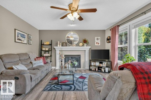 Living room featuring a fireplace with a white mantel, an overhead ceiling fan with lighting, wood-finish flooring, and large windows with privacy blinds - 5322 Terwillegar Boulevard Nw, Edmonton, AB - Indoor Photo Showing Living Room With Fireplace