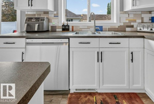 Kitchen featuring white cabinetry with black hardware, dark countertops, an undermount double basin sink with a gooseneck faucet, a built-in dishwasher, and a multi-toned tile backsplash - 5322 Terwillegar Boulevard Nw, Edmonton, AB - Indoor Photo Showing Kitchen With Double Sink