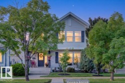 Traditional two-story home featuring light-colored siding, dark window shutters, a prominent front gable, a welcoming front porch, and mature trees in the front yard - 
