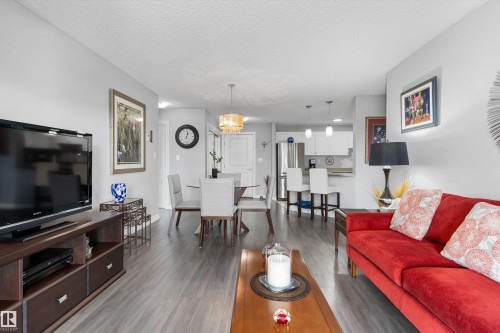 Open-concept living area featuring wood-finish flooring throughout, a red upholstered sofa, dark wood entertainment console with built-in storage, pendant lighting, and a dining area with seating for multiple guests - #305A 6 Spruce Ridge Dr, Spruce Grove, AB - Indoor Photo Showing Living Room