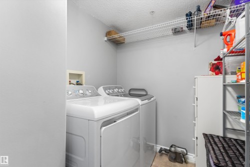 Dedicated laundry room featuring side-by-side washer and dryer with stainless steel accents, overhead wire shelving for storage, white cabinetry, and durable black-finish flooring - #305A 6 Spruce Ridge Dr, Spruce Grove, AB - Indoor Photo Showing Laundry Room