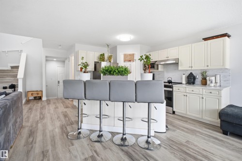 Open-concept kitchen featuring an island with breakfast bar seating, white cabinetry, stainless steel appliances, and wood-finish flooring - 714 87 Street Sw, Edmonton, AB - Indoor Photo Showing Kitchen